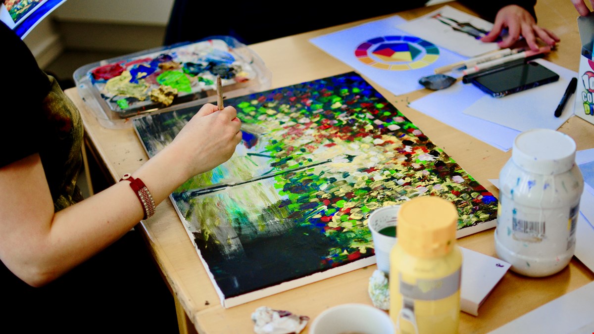 En hand som håller i en pensel och målar en tavla som ligger på ett bord. Tavlan är mycket färgglad.